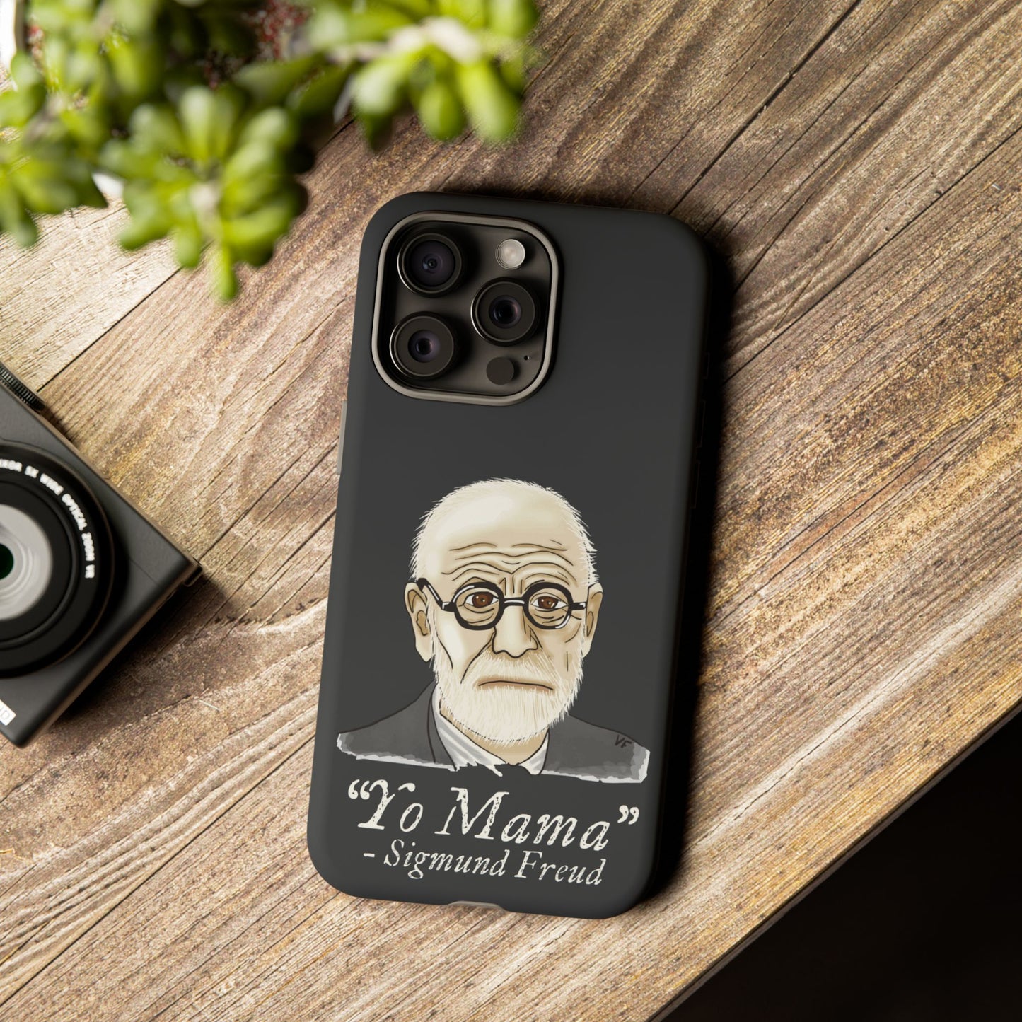 Dark grey phone case featuring a portrait of Sigmund Freud, paired with the cheeky quote “yo mama”—because even the father of psychoanalysis had mommy issues. Here, the case is in use on a phone resting on a table next to a plant and a camera.