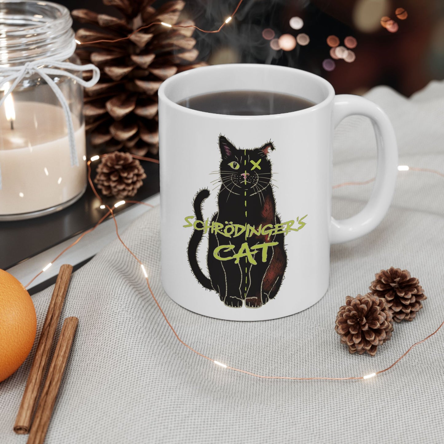 Right view of large white coffee mug featuring a drawing of a black cat that is alive on one half and dead on the other, with the text 'Schrödinger's Cat' printed across the image. The mug is steaming, and sitting next to pine cones and sticks of cinnamon.