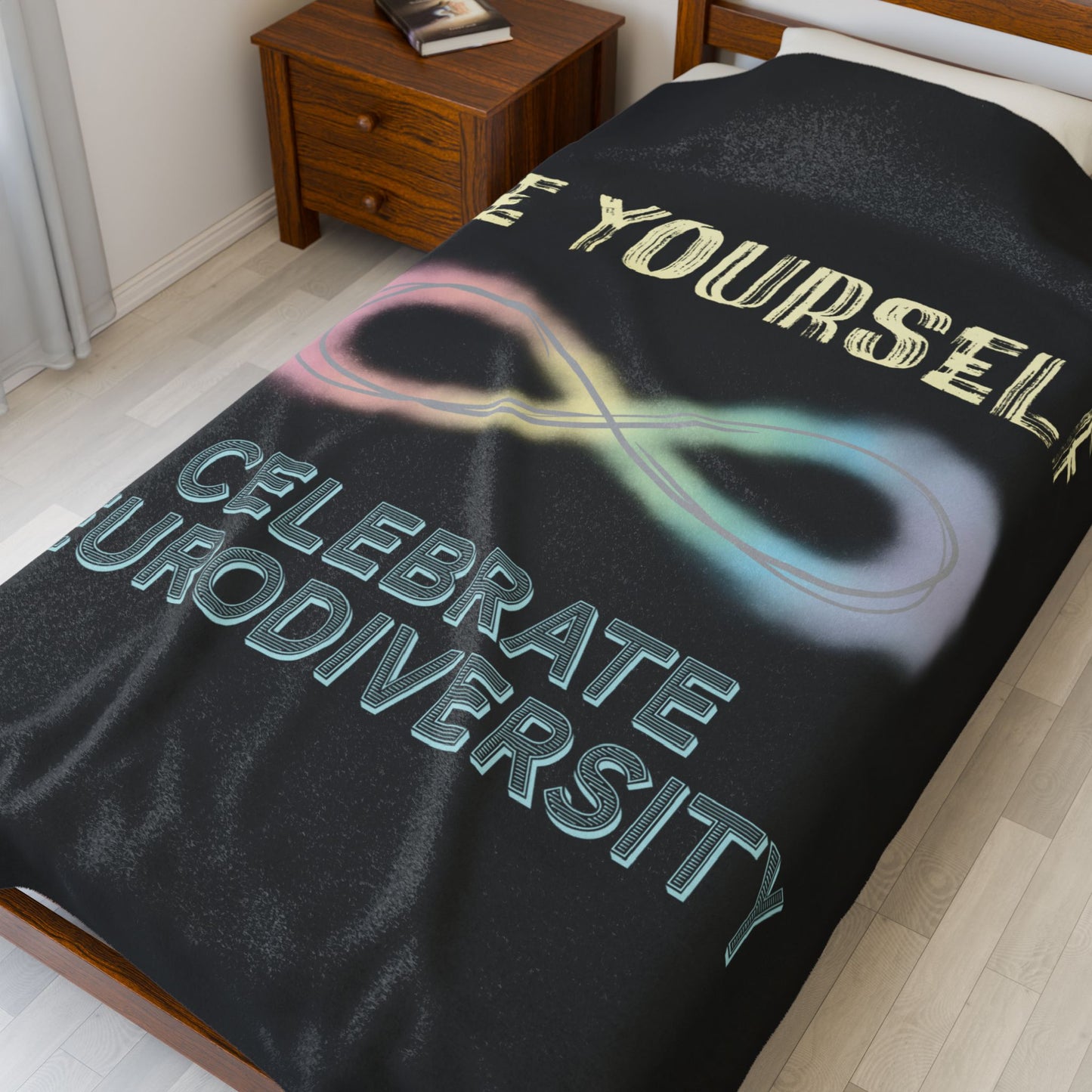 A large black velveteen plush blanket with a pastel rainbow infinity symbol and the uplifting phrase "Be yourself, celebrate neurodiversity"—soft enough to soothe your senses, bold enough to make a statement. Shown on a twin size bed.