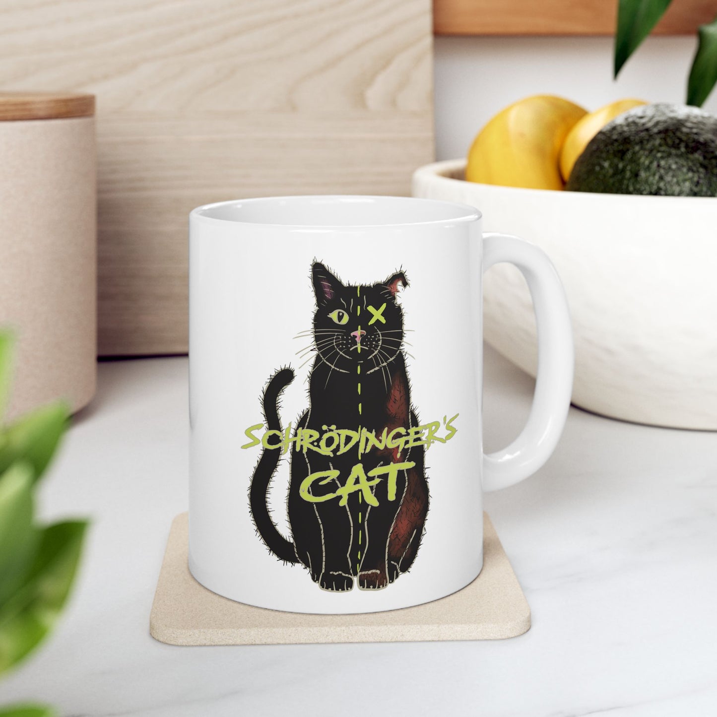 Right view of large white coffee mug featuring a drawing of a black cat that is alive on one half and dead on the other, with the text 'Schrödinger's Cat' printed across the image. The mug is on a coaster on a table next to a bowl of fruit.