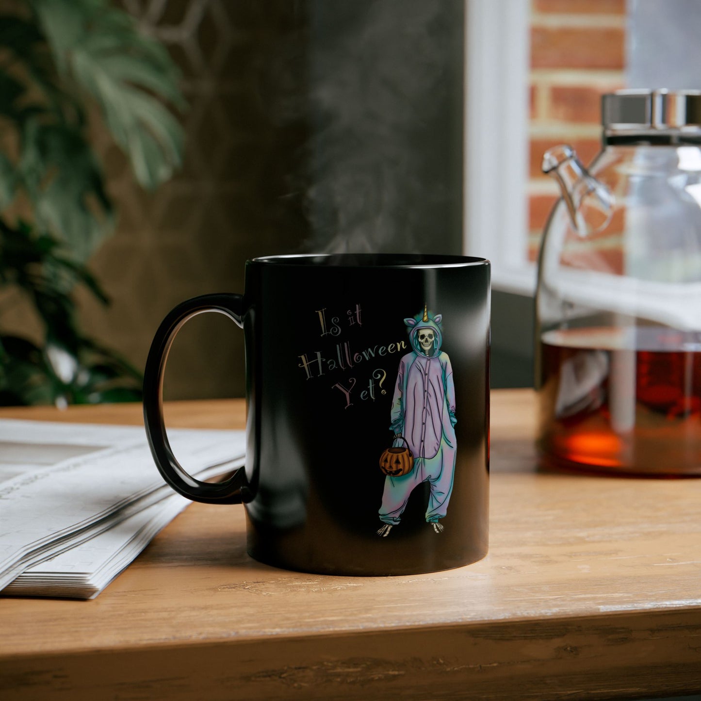 Small black coffee mug sitting on a table featuring a whimsical illustration of a skeleton dressed in a pastel unicorn onesie, complete with a horn and rainbow accents; the playful text reads "Is it Halloween yet?"—because spooky season should be year-round.