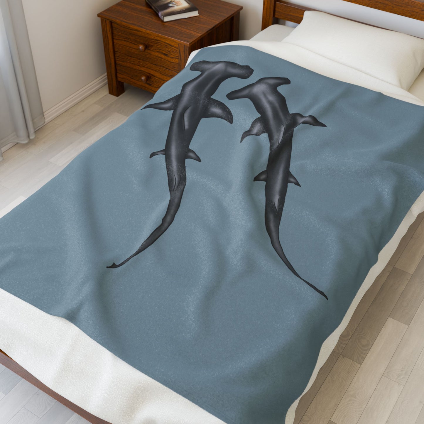 A medium blue-grey velveteen plush blanket featuring two hammerhead sharks gliding through the fabric like it's their personal ocean—perfect for wrapping yourself in comfort with a side of bite. Shown on a twin size bed.