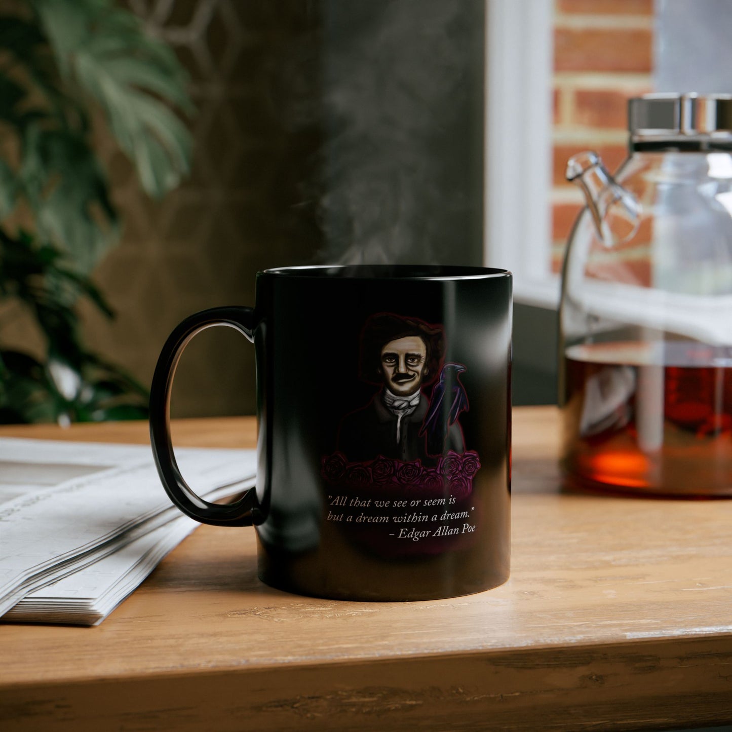 Left view of a small black coffee mug featuring a drawing of Edgar Allan Poe alongside a raven and deep red roses, with one of his famous quotes printed on the side. The mug is sitting on a table near a newspaper and a pot of tea.