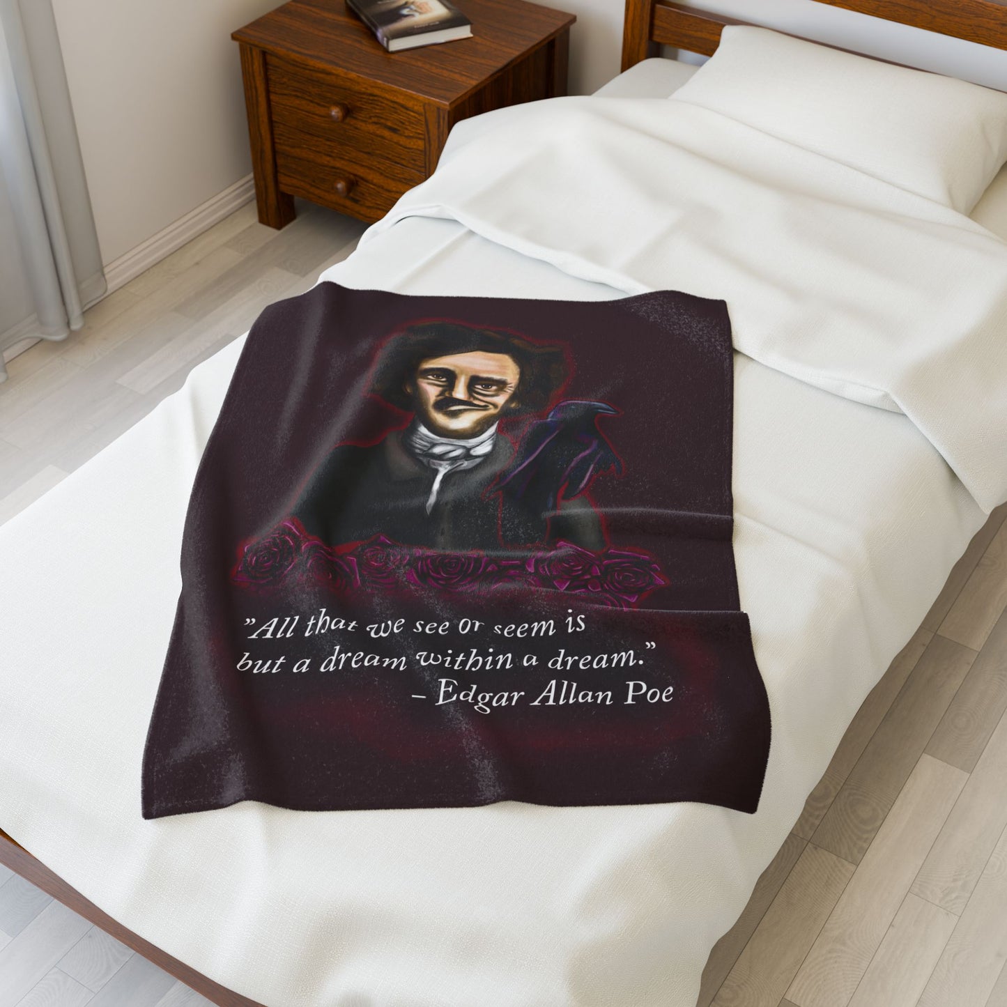 A small burgundy velveteen plush blanket featuring a smirking Edgar Allan Poe, a brooding raven, and crimson roses—perfect for cozying up with your favorite haunting thoughts and the poetic quote, "All that we see or seem is but a dream within a dream. Shown on a twin size bed.
