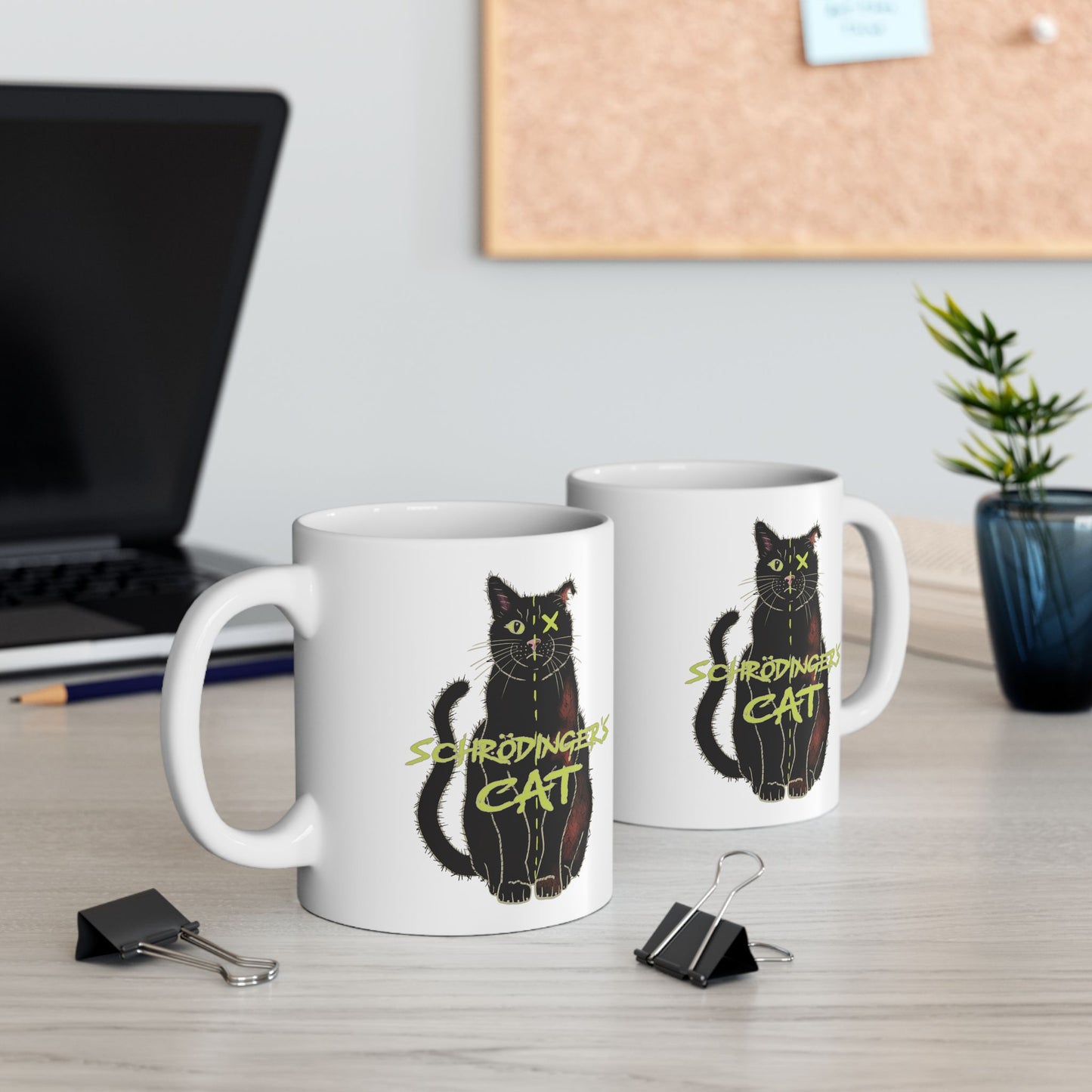 Two white coffee mugs featuring a drawing of a black cat that is alive on one half and dead on the other, with the text 'Schrödinger's Cat' printed across the image. The mugs are on a table next to a laptop, a plant, and other items.