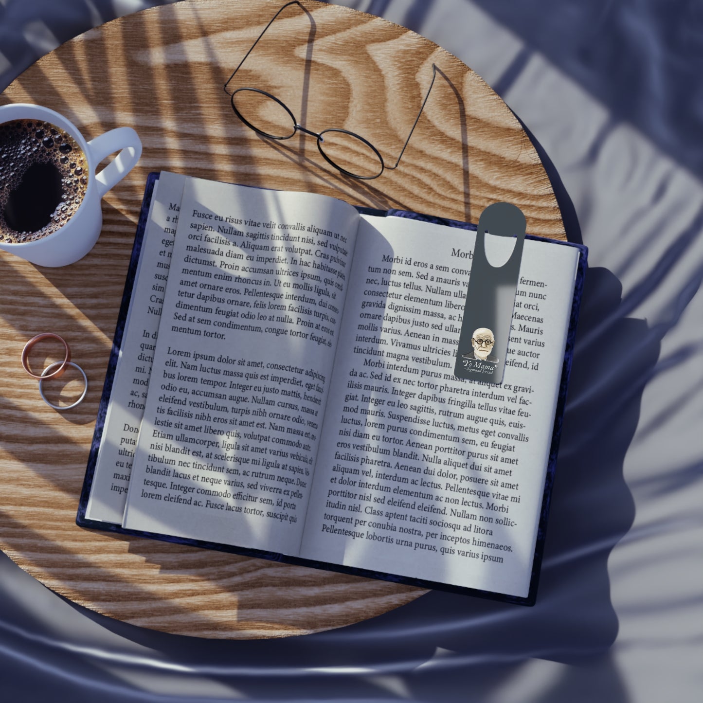 Dark grey-blue aluminum bookmark featuring a portrait of Sigmund Freud with humorous text that reads 'Yo Mama – Sigmund Freud. The bookmark is shown in use holding a page in a book that is resting on a table