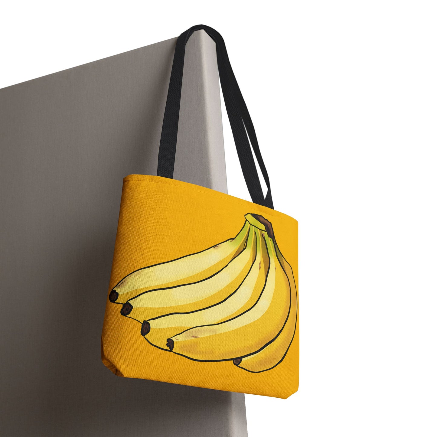 Small yellow tote bag hanging on a chair featuring a playful illustration of a bunch of bananas on the front, set against a solid yellow background.