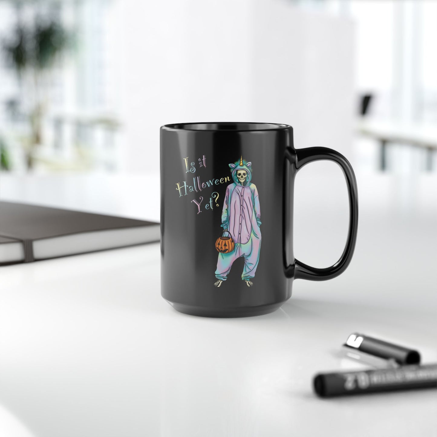 Large black coffee mug sitting on a desk featuring a whimsical illustration of a skeleton dressed in a pastel unicorn onesie, complete with a horn and rainbow accents; the playful text reads "Is it Halloween yet?"—because spooky season should be year-round.