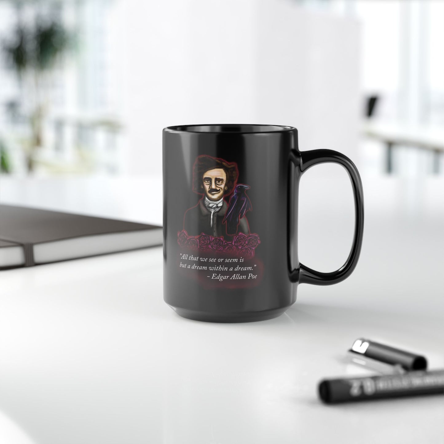 Right view of a small black coffee mug featuring a drawing of Edgar Allan Poe alongside a raven and deep red roses, with one of his famous quotes printed on the side. The mug is sitting on a desk near a notebook and pen.
