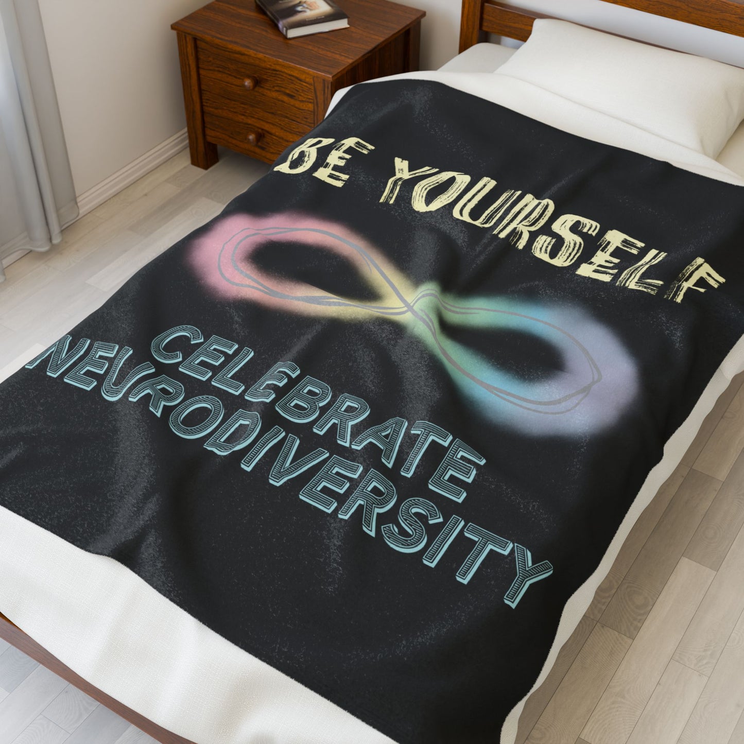 A medium black velveteen plush blanket with a pastel rainbow infinity symbol and the uplifting phrase "Be yourself, celebrate neurodiversity"—soft enough to soothe your senses, bold enough to make a statement. Shown on a twin size bed.