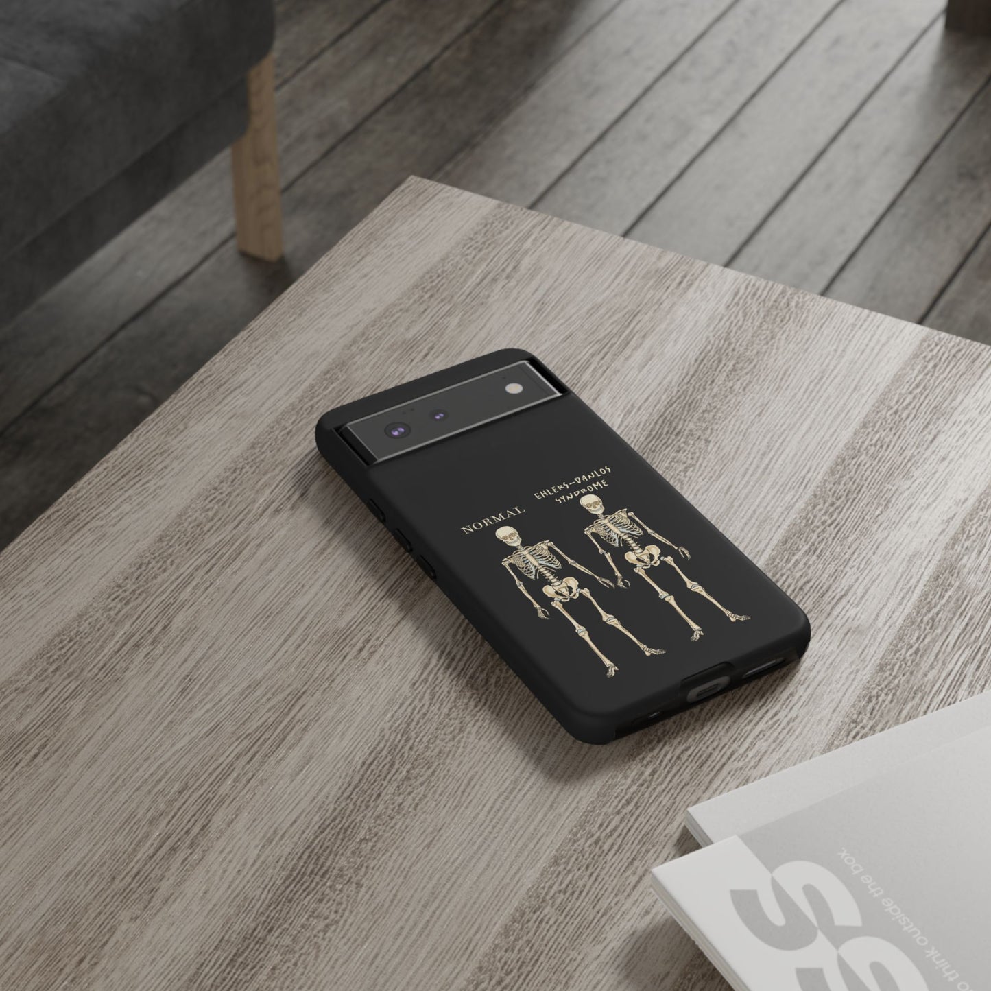 Black phone case with side-by-side skeletons—one labeled “normal,” the other “Ehlers-Danlos Syndrome”—to spread awareness about EDS and its effects. It is shown in use resting on a table.