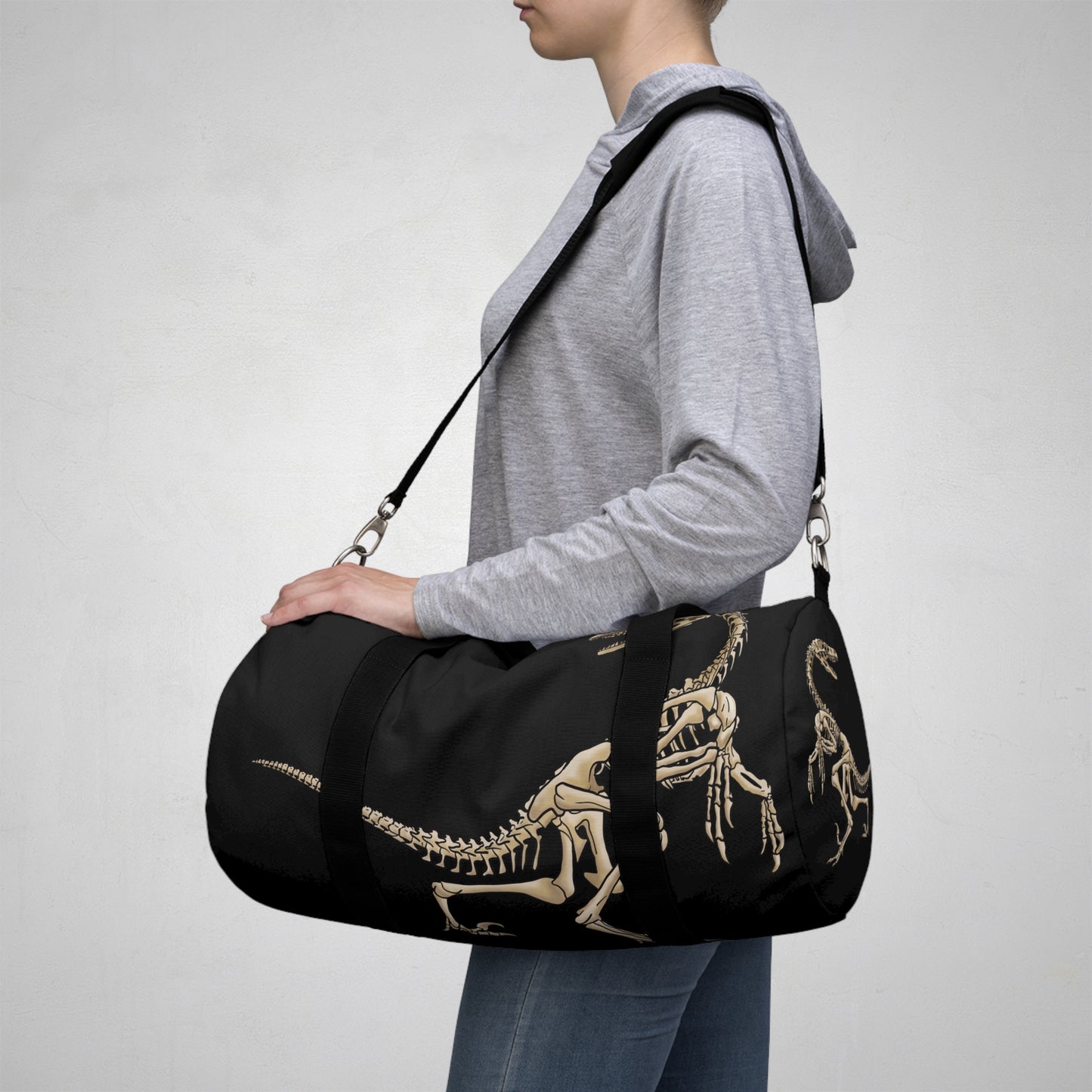 Woman carrying a small black duffel bag with bone-beige illustrations of velociraptor skeleton fossils—perfect for carrying your gear while channeling serious prehistoric predator energy.