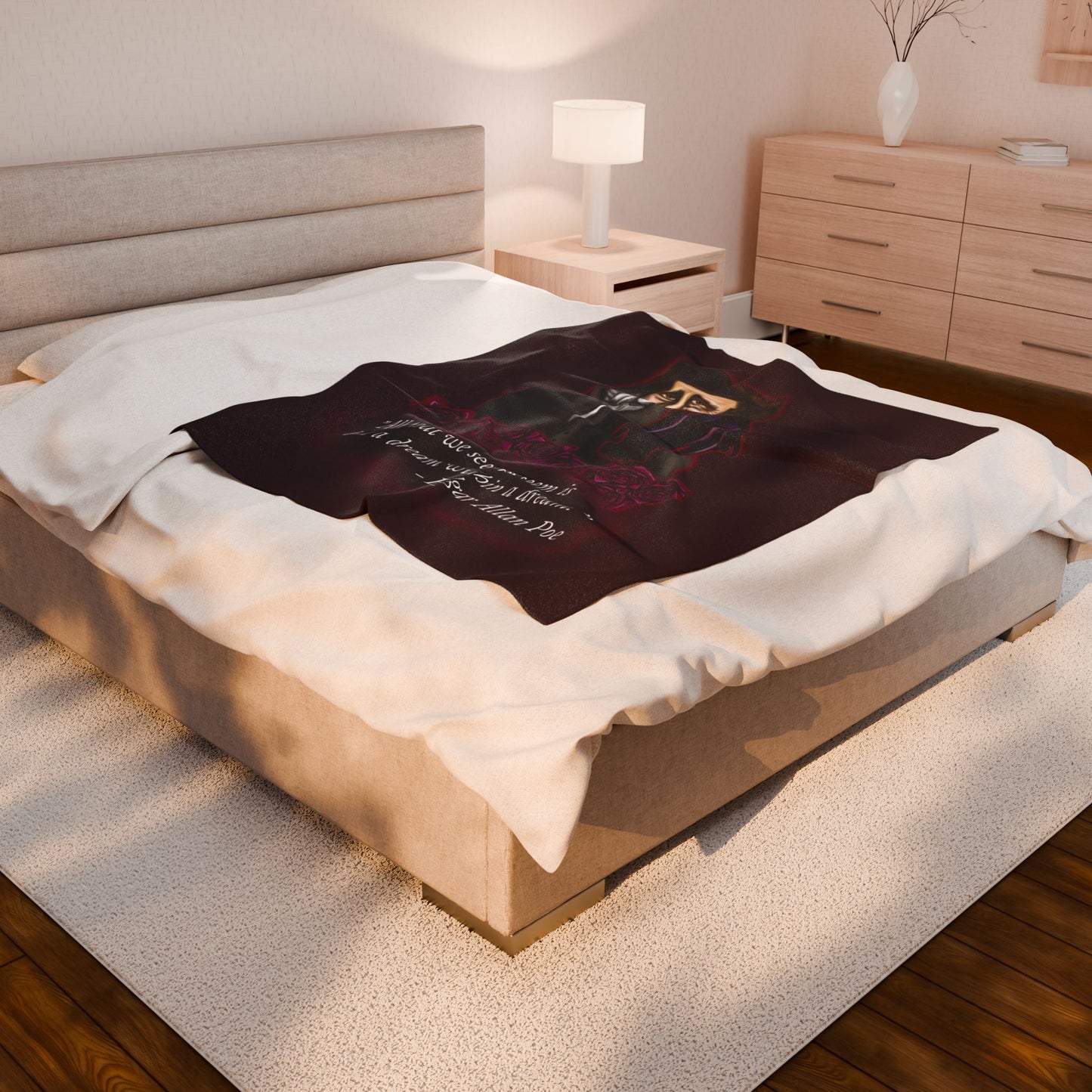 A medium burgundy velveteen plush blanket featuring a smirking Edgar Allan Poe, a brooding raven, and crimson roses—perfect for cozying up with your favorite haunting thoughts and the poetic quote, "All that we see or seem is but a dream within a dream. Shown on a queen size bed.