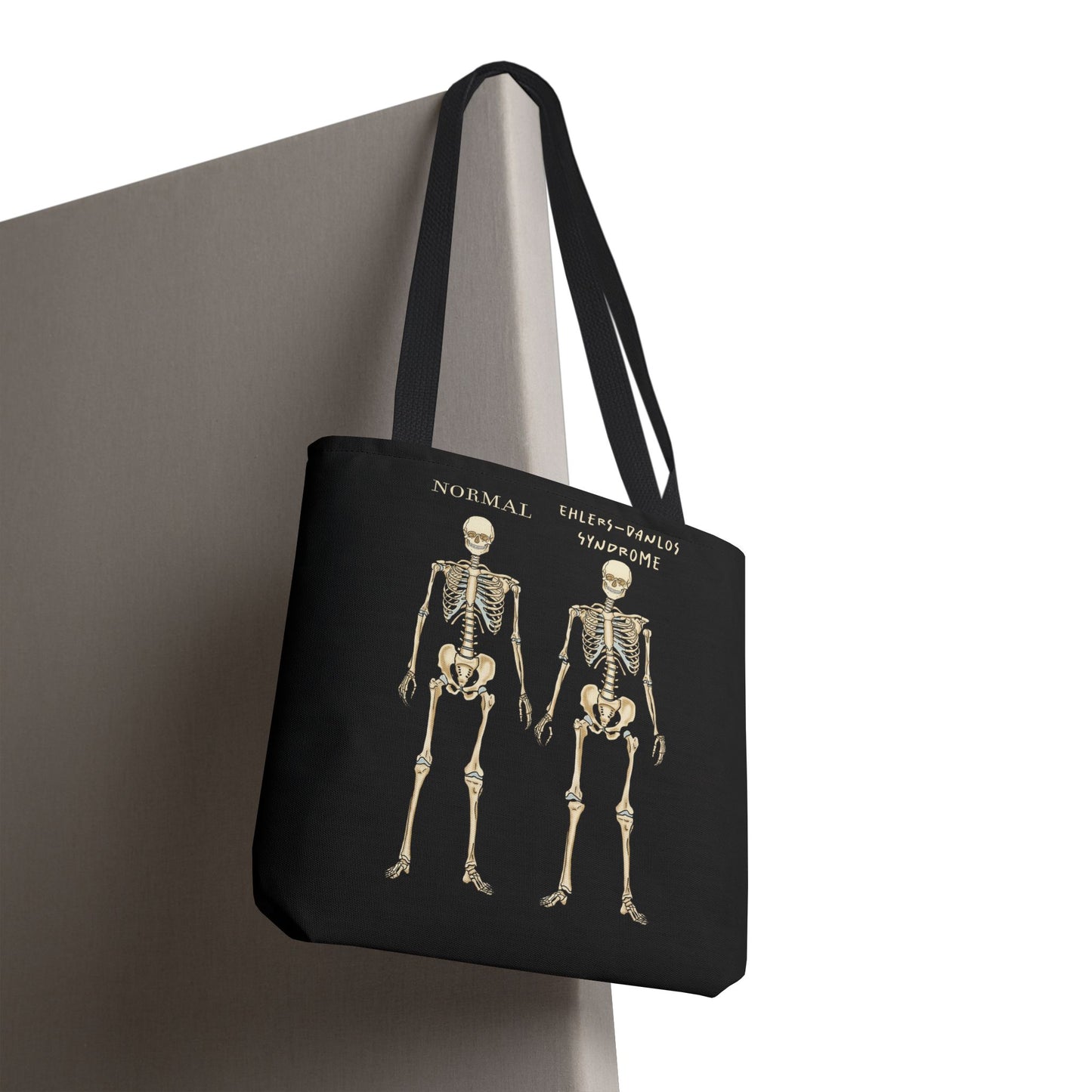 Black tote bag is over a chair. The tote displays two skeletons side by side. The first is labeled 'Normal' and the second is labeled 'Ehlers-Danlos Syndrome,' visually illustrating the skeletal effects of EDS.