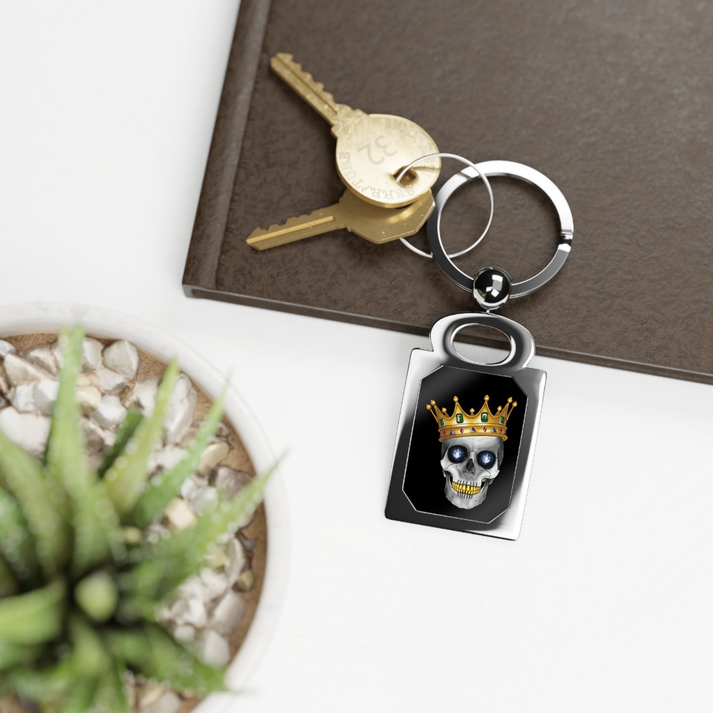 Rectangular silver keychain resting on a table with a bold design framed inside: a skull with gleaming gold teeth, glittering sapphire eyes, and a lavish gold crown—all set against a dramatic black background, serving royal attitude with a side of macabre flair.