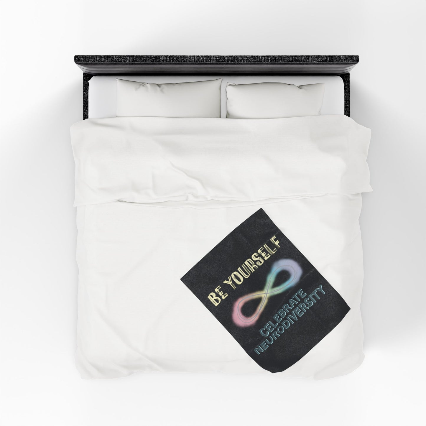 A small black velveteen plush blanket with a pastel rainbow infinity symbol and the uplifting phrase "Be yourself, celebrate neurodiversity"—soft enough to soothe your senses, bold enough to make a statement. Shown on a queen size bed.