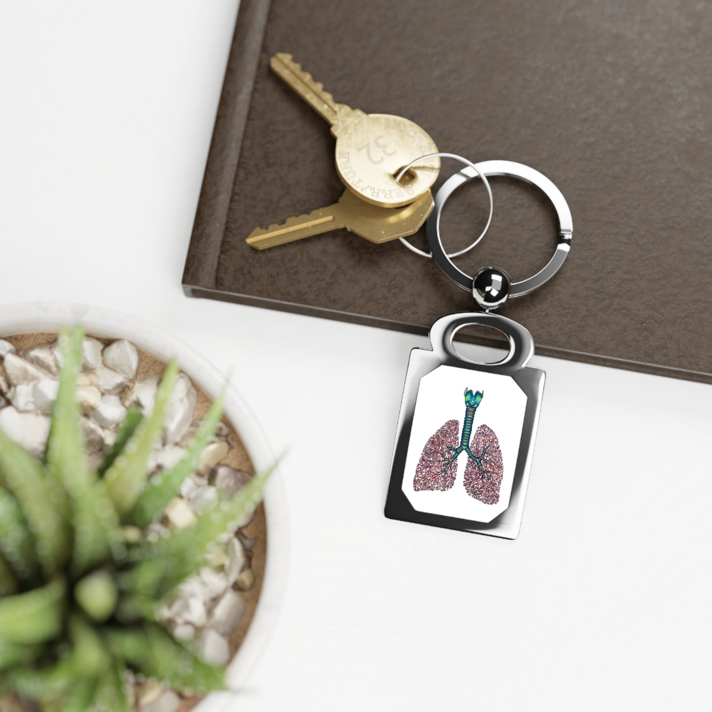 Rectangular silver frame keychain featuring a detailed anatomical drawing of lungs, including the bronchial tree, trachea, and larynx, set against a white background is sitting on top of a book on a white table, next to a plant.