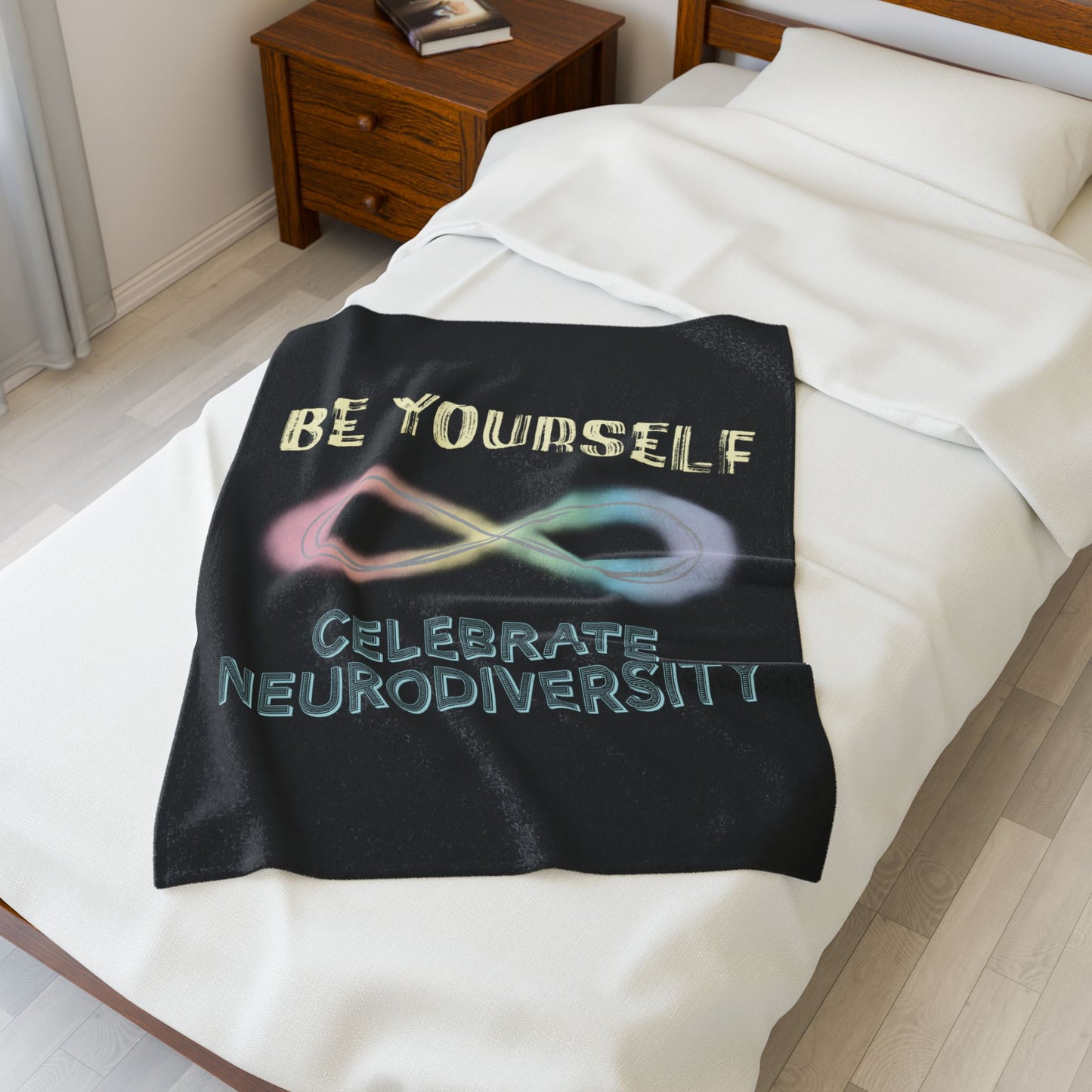 A small black velveteen plush blanket with a pastel rainbow infinity symbol and the uplifting phrase "Be yourself, celebrate neurodiversity"—soft enough to soothe your senses, bold enough to make a statement. Shown on a twin size bed.