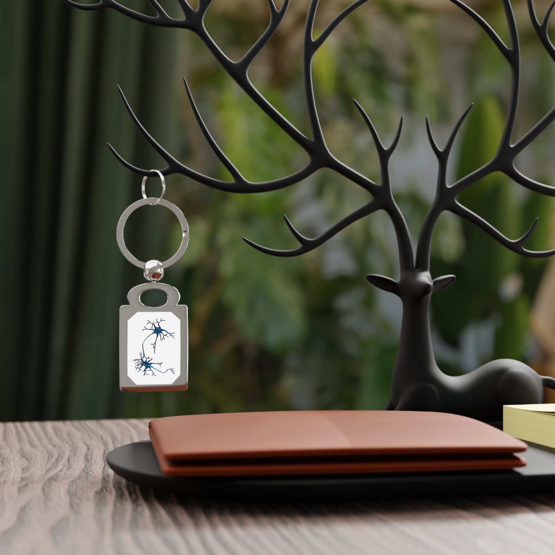A rectangular silver frame keychain featuring a drawing of two blue neurons forming a synapse, set against a white background, is hanging on display.