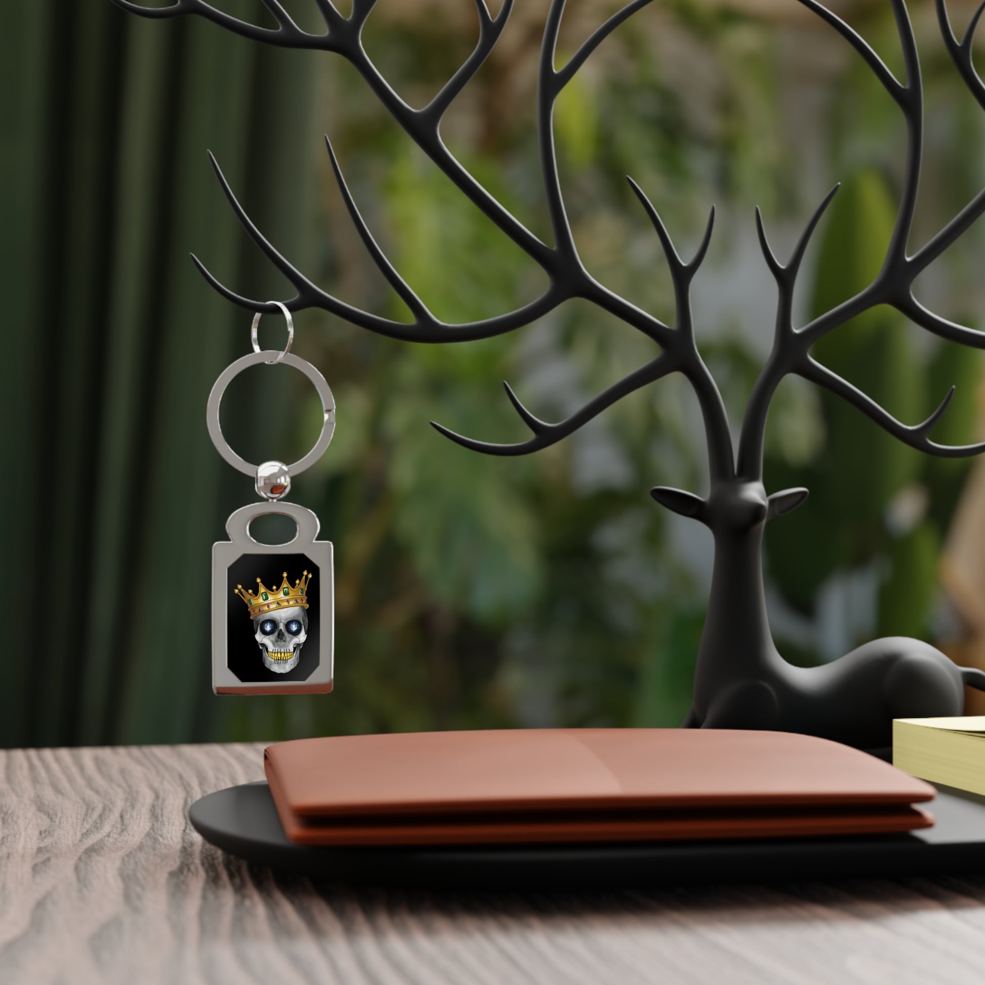Hanging on display, there is a rectangular silver keychain with a bold design framed inside: a skull with gleaming gold teeth, glittering sapphire eyes, and a lavish gold crown—all set against a dramatic black background, serving royal attitude with a side of macabre flair.