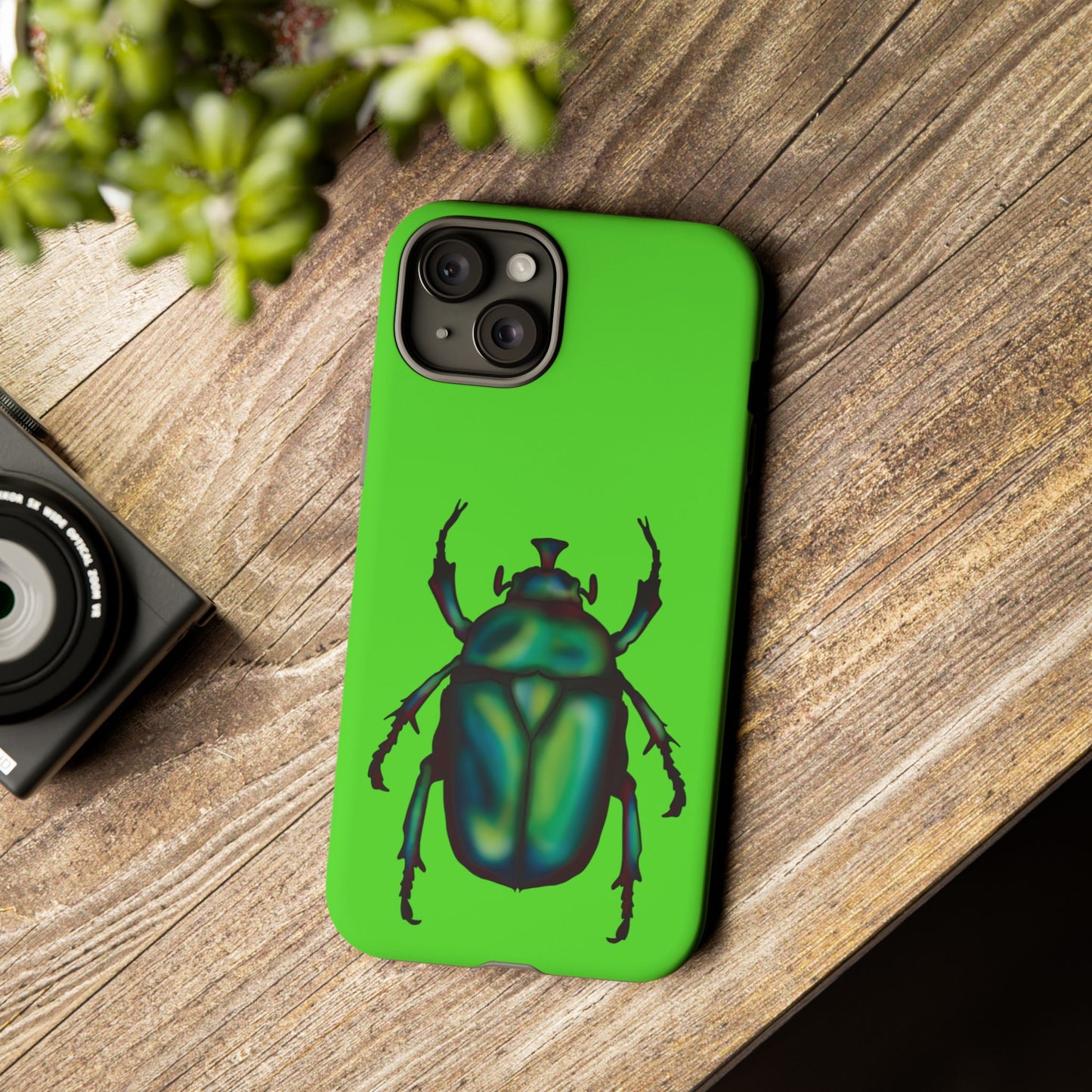 Lime green phone case featuring a dazzling illustration of a green and blue June bug—this iridescent little beetle brings all the shine without the buzz. It is shown sitting on a table next to a plant and a camera.