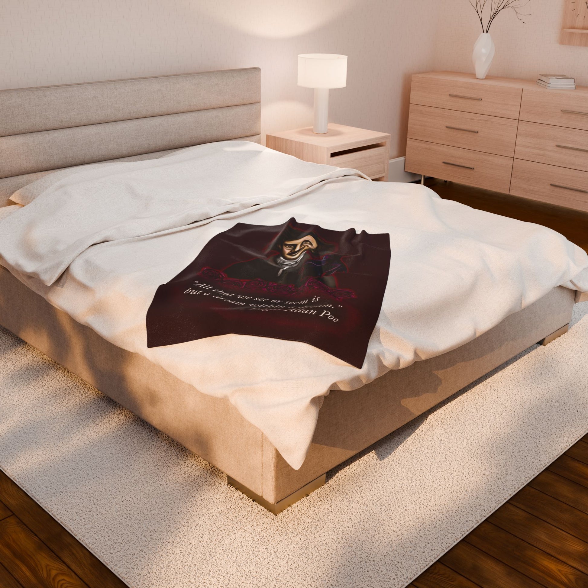 A small burgundy velveteen plush blanket featuring a smirking Edgar Allan Poe, a brooding raven, and crimson roses—perfect for cozying up with your favorite haunting thoughts and the poetic quote, "All that we see or seem is but a dream within a dream. Shown on a queen size bed.