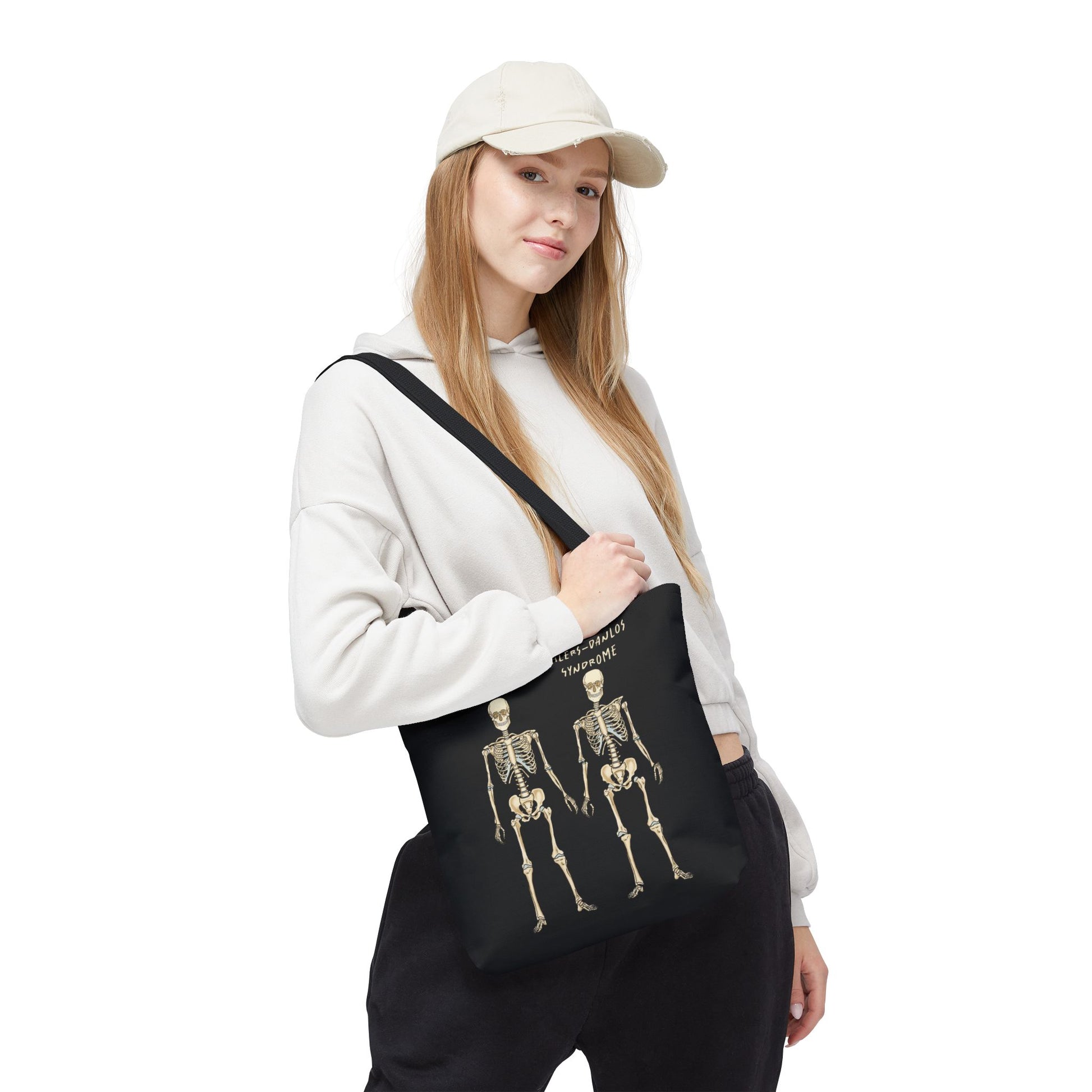 Small black tote bag is over a woman's shoulder. The tote displays two skeletons side by side. The first is labeled 'Normal' and the second is labeled 'Ehlers-Danlos Syndrome,' visually illustrating the skeletal effects of EDS.