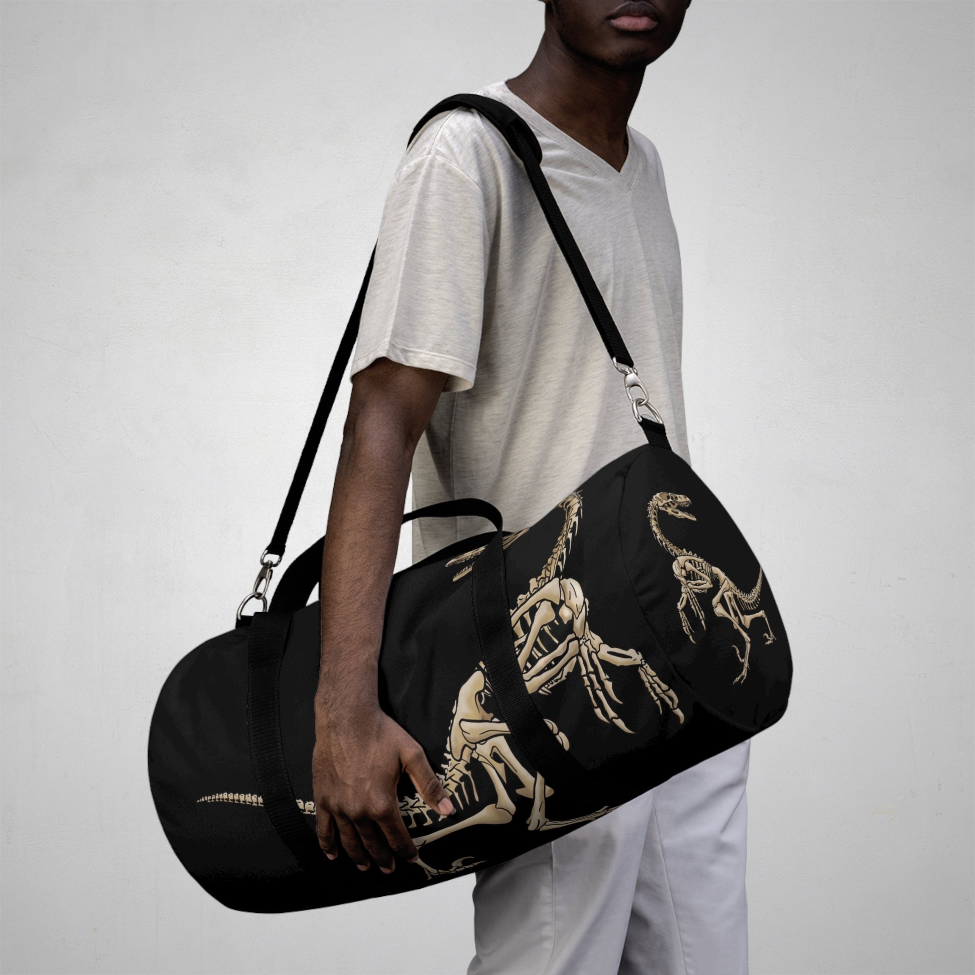 Man carrying a large black duffel bag with bone-beige illustrations of velociraptor skeleton fossils—perfect for carrying your gear while channeling serious prehistoric predator energy.