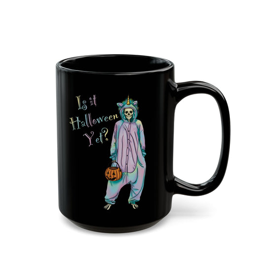 Black coffee mug featuring a whimsical illustration of a skeleton dressed in a pastel unicorn onesie, complete with a horn and rainbow accents; the playful text reads "Is it Halloween yet?"—because spooky season should be year-round.