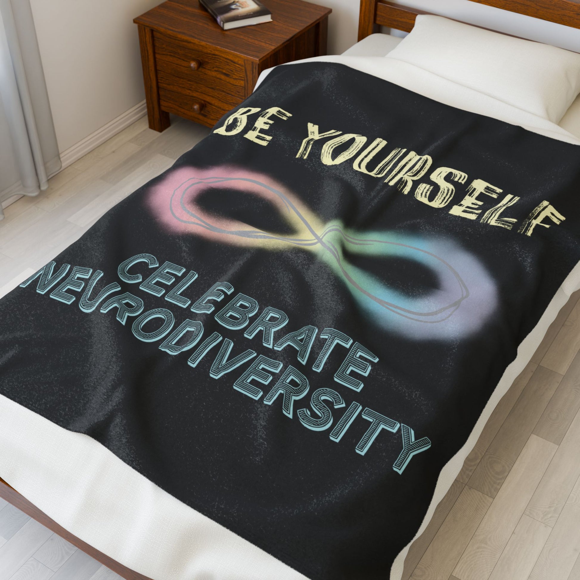 A medium black velveteen plush blanket with a pastel rainbow infinity symbol and the uplifting phrase "Be yourself, celebrate neurodiversity"—soft enough to soothe your senses, bold enough to make a statement. Shown on a twin size bed.