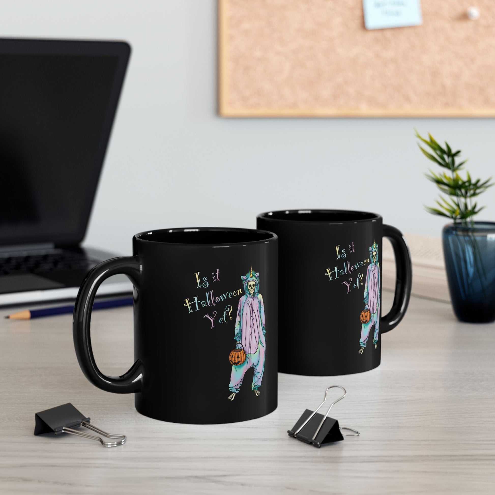 Two black coffee mugs sitting on a desk featuring a whimsical illustration of a skeleton dressed in a pastel unicorn onesie, complete with a horn and rainbow accents; the playful text reads "Is it Halloween yet?"—because spooky season should be year-round.