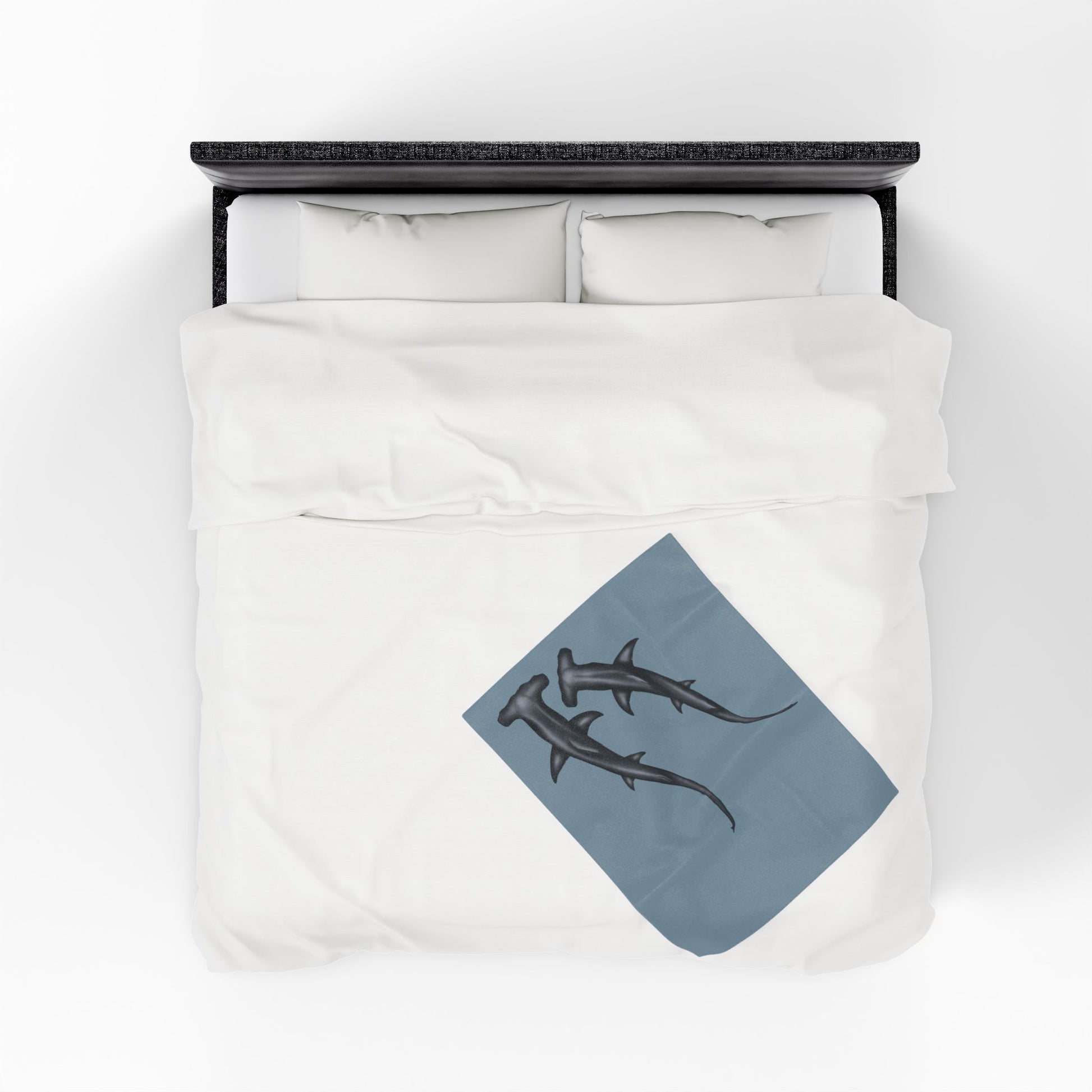 A small blue-grey velveteen plush blanket featuring two hammerhead sharks gliding through the fabric like it's their personal ocean—perfect for wrapping yourself in comfort with a side of bite. Shown on a queen size bed from above.