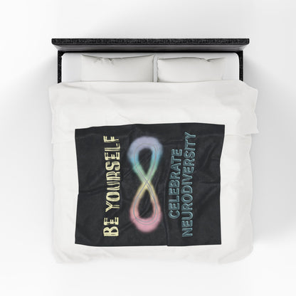 A medium black velveteen plush blanket with a pastel rainbow infinity symbol and the uplifting phrase "Be yourself, celebrate neurodiversity"—soft enough to soothe your senses, bold enough to make a statement. Shown on a queen size bed.