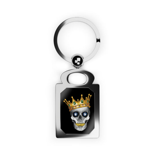 Rectangular silver keychain with a bold design framed inside: a skull with gleaming gold teeth, glittering sapphire eyes, and a lavish gold crown—all set against a dramatic black background, serving royal attitude with a side of macabre flair.