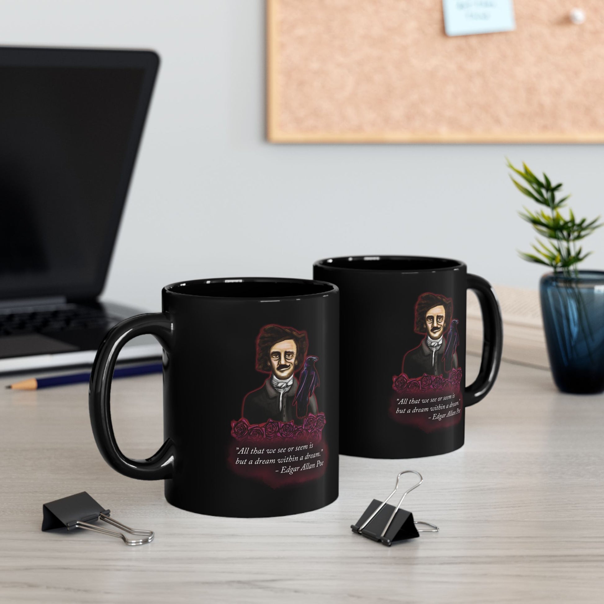 Two black coffee mugs featuring a drawing of Edgar Allan Poe alongside a raven and deep red roses, with one of his famous quotes printed on the side. The mugs are sitting on a desk near a computer and a plant.