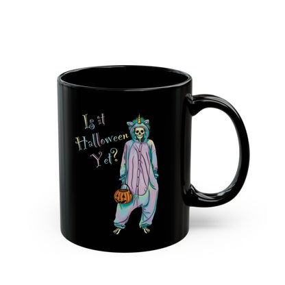 Right view of a small black coffee mug featuring a whimsical illustration of a skeleton dressed in a pastel unicorn onesie, complete with a horn and rainbow accents; the playful text reads "Is it Halloween yet?"—because spooky season should be year-round.
